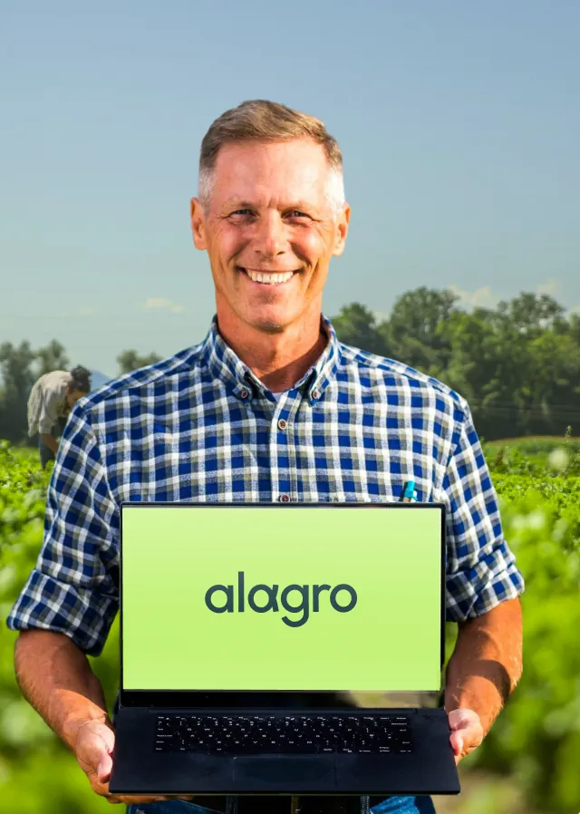 Productor sonriendo en un campo de papas, con una notebook que muestra el logo de Alagro en la pantalla